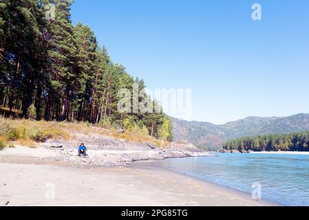 An elderly woman sits on a stone on the beach of the mountain river Katun in autumn in Altai. Stock Photo