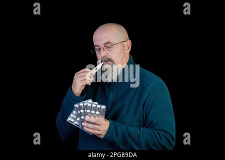 A middle-aged man checking his temperature with a bunch of pills in his hands, isolated on black background Stock Photo