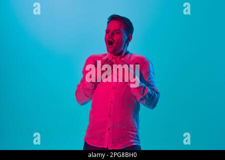 Portrait of young emotional man in white shirt posing with shocked expression against blue studio background in pink neon light. Stock Photo