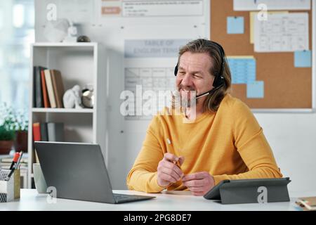 Mature teacher in headphones sitting at his workplace with laptop and talking to his students online Stock Photo