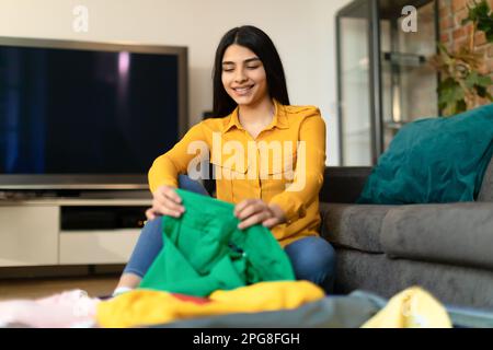 Young spanish lady packing suitcase for vacation, sitting near suitcase, preparing for travel in living room at home Stock Photo
