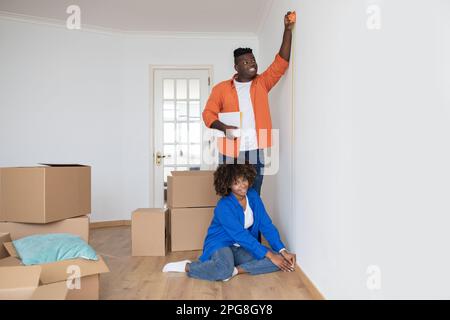 Young Black Spouses Using Tape Ruler, Taking Measurements In Living Room Together Stock Photo