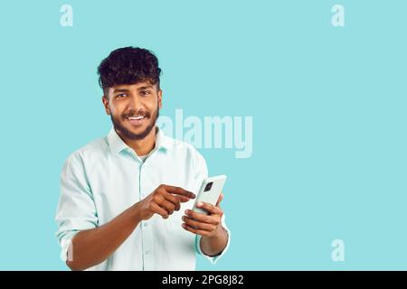 Young satisfied Indian man smiling holding phone in hand and looking at camera stands in studio Stock Photo