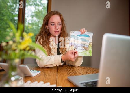 A young art teacher holding a drawing that she made and explaining complicated techniques to the students during an online class Stock Photo