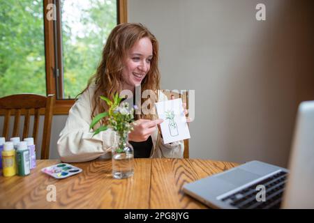 A young art teacher holding a drawing that she made and explaining complicated techniques to the students during an online class Stock Photo