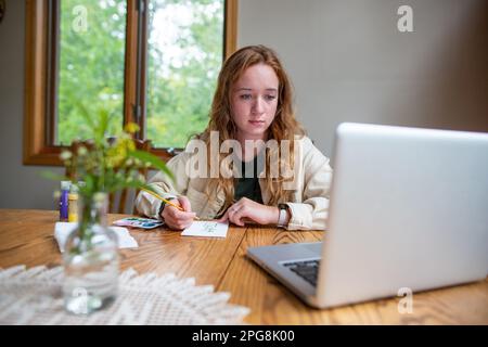 A young woman looking at her laptop and carefully drawing something on a piece of paper Stock Photo