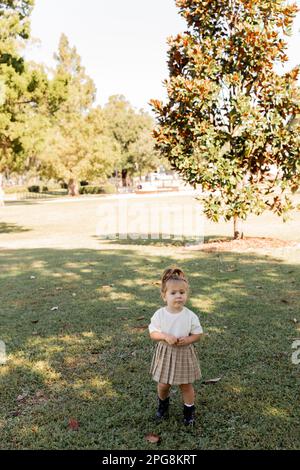 full length of toddler girl in skirt and white t-shirt standing in park with green trees,stock image Stock Photo