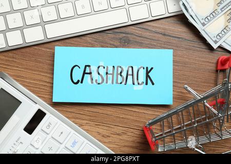 Card with word Cashback, calculator, keyboard, shopping cart and dollar banknotes on wooden background, flat lay Stock Photo