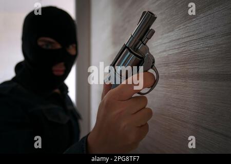 Terrorist hooded in balaclava, holding a gun in hand. Man aiming with a pistol, dark building indoor background, Killer, armed robbery concept. Stock Photo