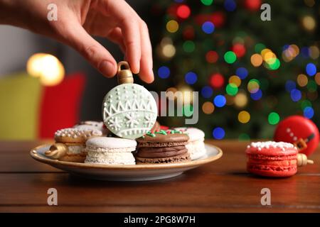 Woman taking Christmas macaron from plate at wooden table against blurred festive lights, closeup Stock Photo