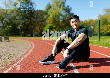 Young asian sportsman, spotter, runner sitting in stadium on treadmill and resting after jogging, exercise. Tired but smiling, he looks at the camera. Stock Photo