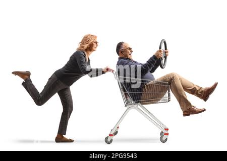 Woman pushing a mature man inside a shopping cart sitting and holding a steering wheel isolated on white background Stock Photo