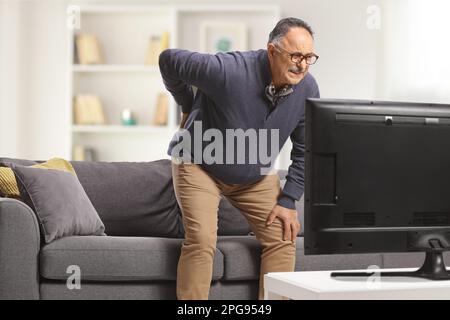 Mature man with back pain trying to sit on a sofa in front of tv Stock Photo