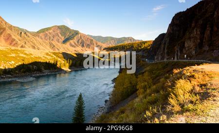 The mountain road Chuisky tract goes under a rock in the shade next to the Katun River in Altai in the evening in Siberia in autumn. Stock Photo
