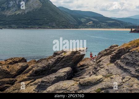 two young women and a man sunbathing in swimsuits on the rocks of the town of Sonabia, the Cantabrian Sea and the mountains in the background, Spa Stock Photo