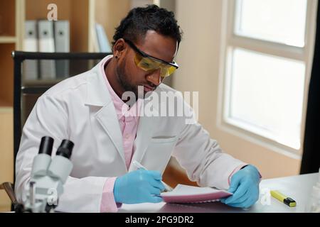 Young man in lab coat and gloves making notes in copybook while sitting by workplace and carrying out new scientific investigation Stock Photo