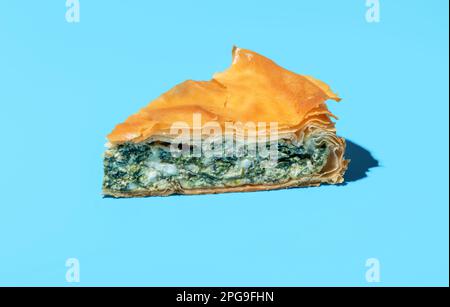 Close-up with a single slice of phyllo cake in bright light on a blue table. Homemade savory pie with feta cheese and green spinach. Stock Photo