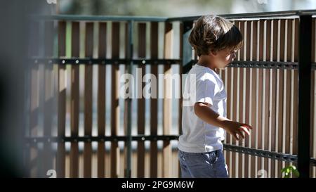 Child standing by balcony fence looking outdoors from second floor. Child holding in wooden bars staring outside Stock Photo