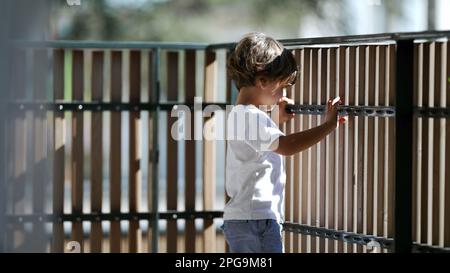 Child standing by balcony fence looking outdoors from second floor. Child holding in wooden bars staring outside Stock Photo