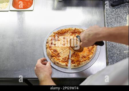 Chef cutting freshly baked pizza with round knife on wooden board Stock Photo