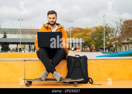 Stylish bearded casual young man freelancer working with a modern laptop and using in a smartphone while sitting on a bench with a longboard outdoors Stock Photo