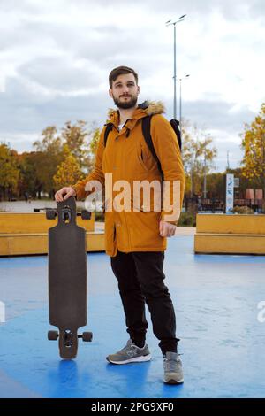 Full-height Portrait of stylish bearded casual young man in a yellow jacket with a longboard standing at the skating park outdoors Stock Photo