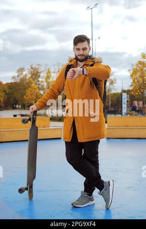 Full-height Portrait of stylish bearded casual young man in a yellow jacket with a longboard standing at the skating park watch the clock outdoors Stock Photo