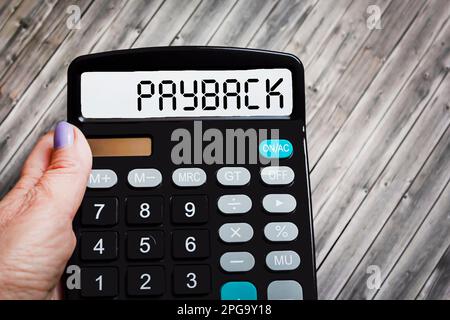 The word PAYBACK is written on a calculator in a girl's hand on a wooden table. Stock Photo