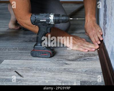 a man installs a floor wooden plinth made of dark lacquered wood in a room with gray textured linoleum using a screwdriver, holding the plank Stock Photo