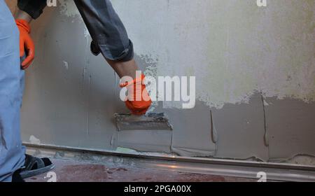 a plasterer in orange gloves applies a viscous elastic mortar with a wide spatula on a room wall, texture stains of putty on the wall Stock Photo