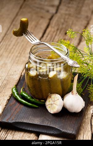 Pickled gherkins in a transparent glass jar, one canned cucumber on a vintage fork, fresh garlic, herbs and spices on a wooden background. Stock Photo