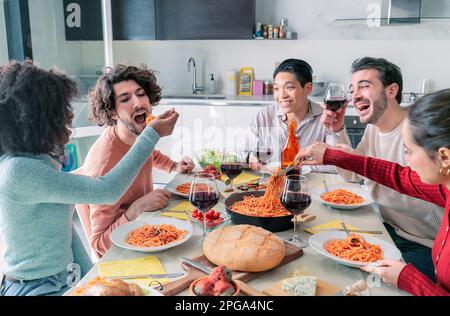 Happy young ethnic woman spoon feeding male friend with delicious pasta in kitchen. Friends eating together Stock Photo