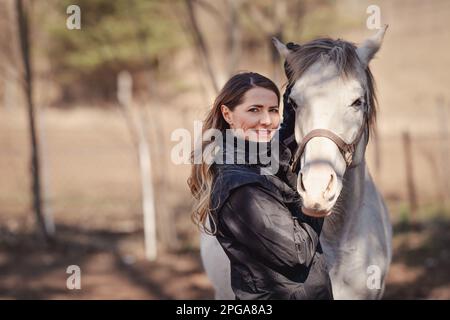 Young woman standing next to white Arabian horse, holding it by ears, sun shines on them, closeup detail Stock Photo