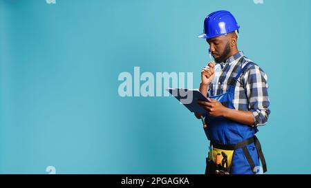 Confident builder making calculations on papers, taking notes on files before starting professional construction project in studio. Male carpenter writing information and measurements. Stock Photo