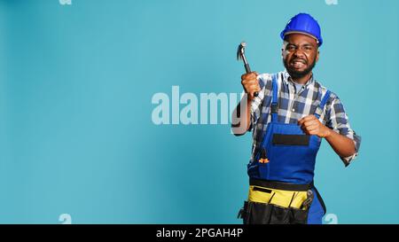 Construction worker hitting something with hammer, using manual metallic tool for renovation and building job. Young builder in uniform and hardhat holding mullet instrument on camera. Stock Photo