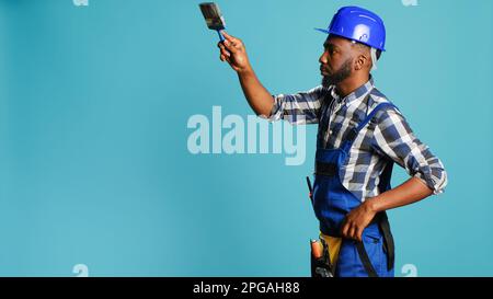 Young carpenter painting walls with paintbrush on camera, using renovating tools to change color. Professional craftsman working with brush on reconstruction equipment on blue backdrop. Stock Photo