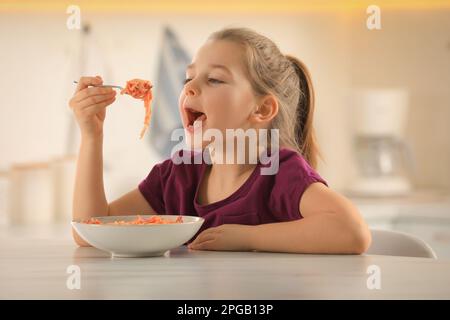 Cute little girl eating tasty pasta at table in kitchen Stock Photo