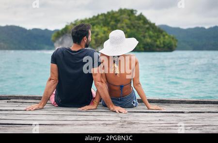 The best vacations are the ones with him. Rearview shot of an unrecognizable couple sitting together on a boardwalk overlooking the ocean during Stock Photo