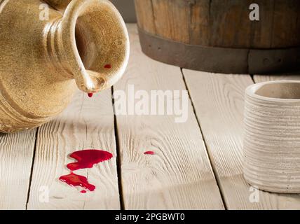 Clay wine jug, wine barrel and clay glass on a wooden table. A puddle of spilled red wine under a jug Stock Photo
