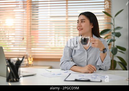 Relaxed and happy young Asian businesswoman sipping morning coffee, looking aside, daydreaming, resting at her desk in her office. Stock Photo
