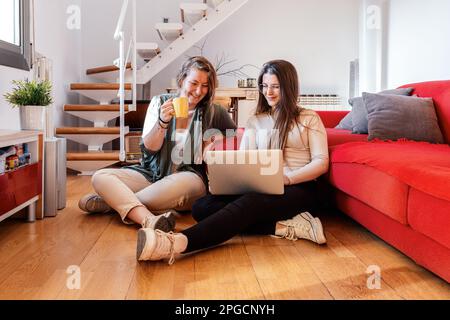 Positive young female friends relaxing on floor near comfortable sofa while one enjoying coffee and together looking at screen of laptop during search Stock Photo