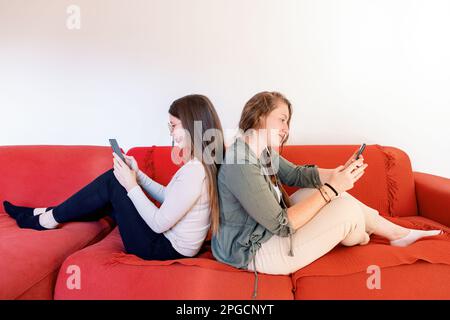 Side view of young female friends relaxing on sofa and sitting with backs against each other with raised legs while looking at screen of smartphones i Stock Photo