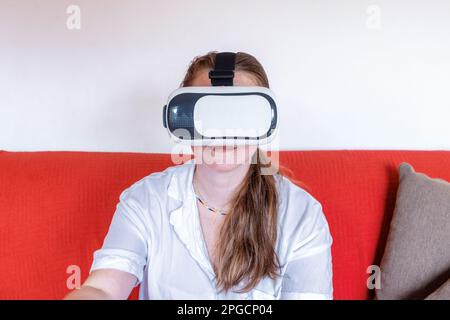Positive young female using virtual reality headset goggles and looking at screen while watching improved entertaining cyberspace and relaxing on sofa Stock Photo