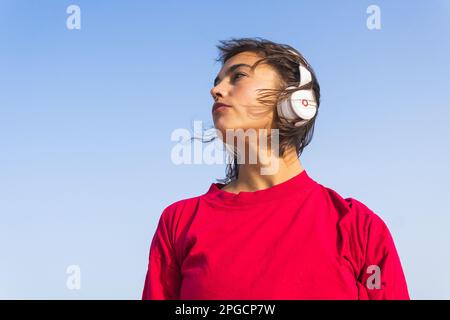 From below thoughtful young female in red t shirt looking away listening to music in headphones while resting near sea Stock Photo