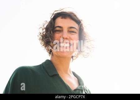Low angle of happy young female with curly short hair and piercing smiling happily while looking at camera against bright light Stock Photo