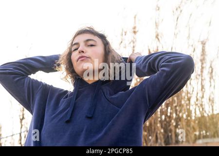 Low angle of calm young female with wavy hair and piercing wearing hoodie looking at camera on street Stock Photo