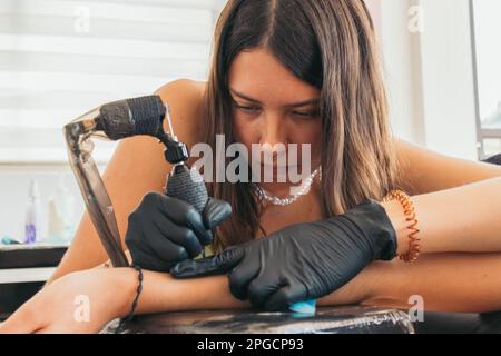 Young female master in gloves working with tattoo machine while painting artwork on anonymous female in salon Stock Photo