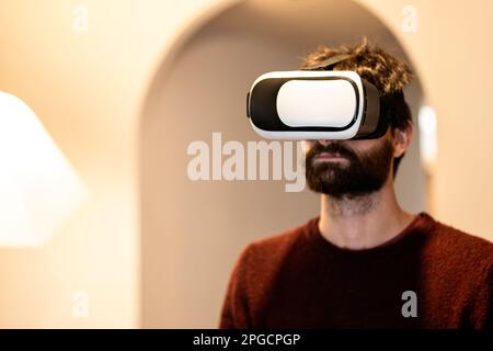 Confident bearded male in casual clothes experiencing virtual reality using goggles against wall Stock Photo