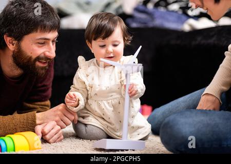 Cheerful bearded father looking at adorable little daughter playing with wind turbine toy sitting near crop mother at home Stock Photo