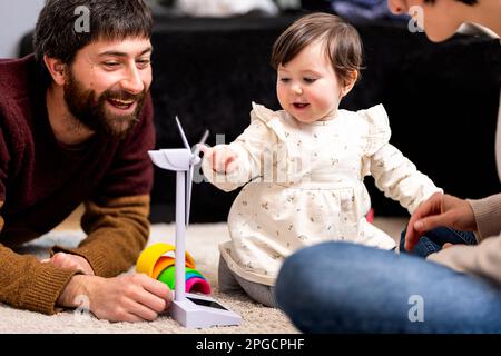 Cheerful bearded father looking at adorable little daughter playing with wind turbine toy sitting near crop mother at home Stock Photo